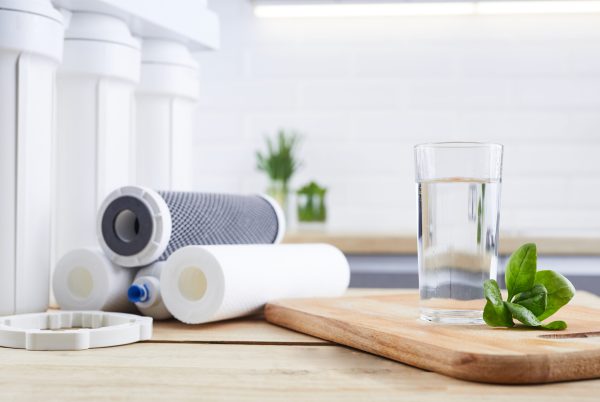 A glass of clean water with osmosis filter, green leaves and cartridges on wooden table in kitchen interior. Concept Household filtration system.